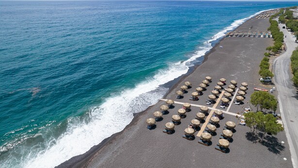 Vista aérea de una playa con hileras de sombrillas de paja y un paseo pavimentado a lo largo de la orilla.