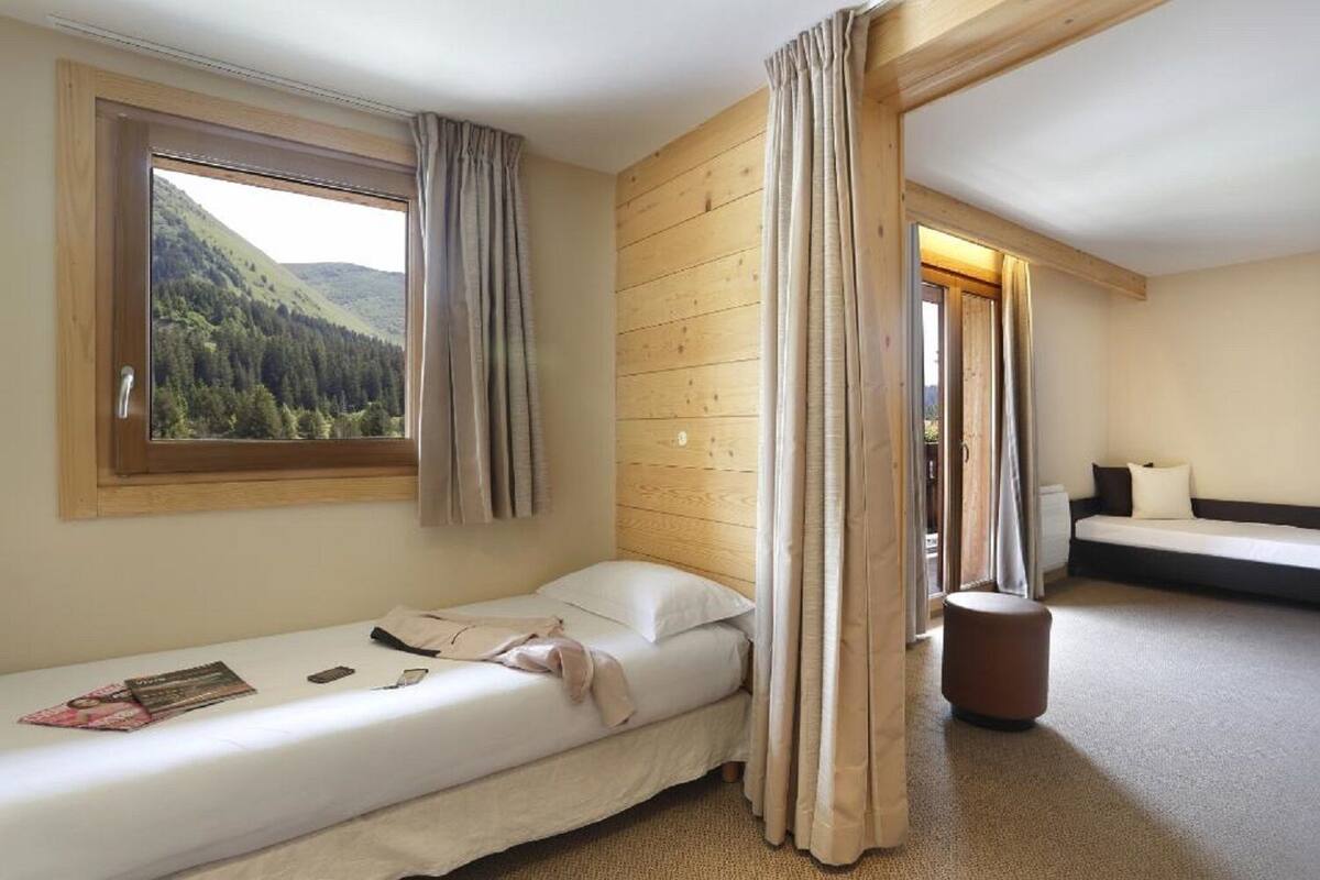 Habitació petita amb un llit individual, sòl de fusta, finestra amb cortines i vista a una vessant boscosa.