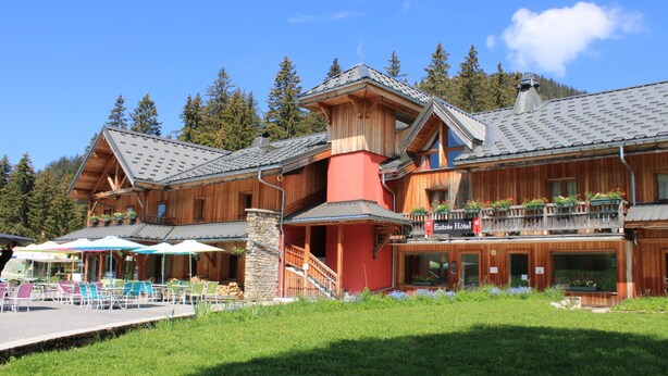 A wooden chalet with a stone staircase, outdoor seating, and a sign for 'Entrée Hotel'.