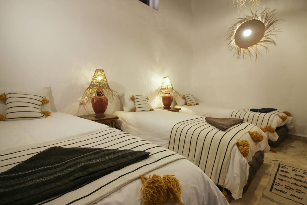 Una habitación de hotel con tres camas, cada una con ropa de cama a rayas y almohadas decorativas. Hay lámparas de pared a cada lado de las camas.