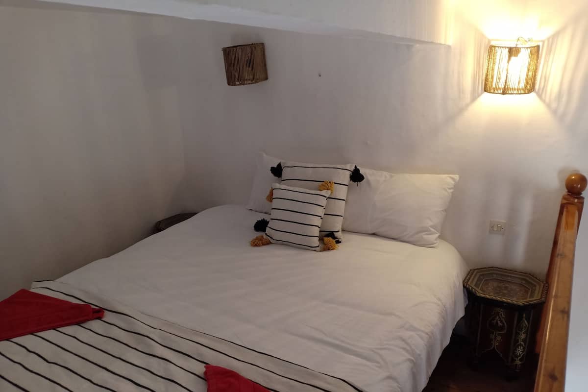 Una cama bien hecha con ropa de cama a rayas y dos almohadas, un taburete de madera pequeño y una lámpara de pared.