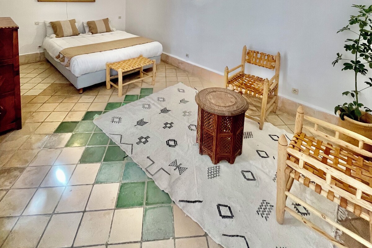 Une pièce comprenant un lit, une chaise en bois, une petite table, une plante en pot et un tableau accroché au mur.