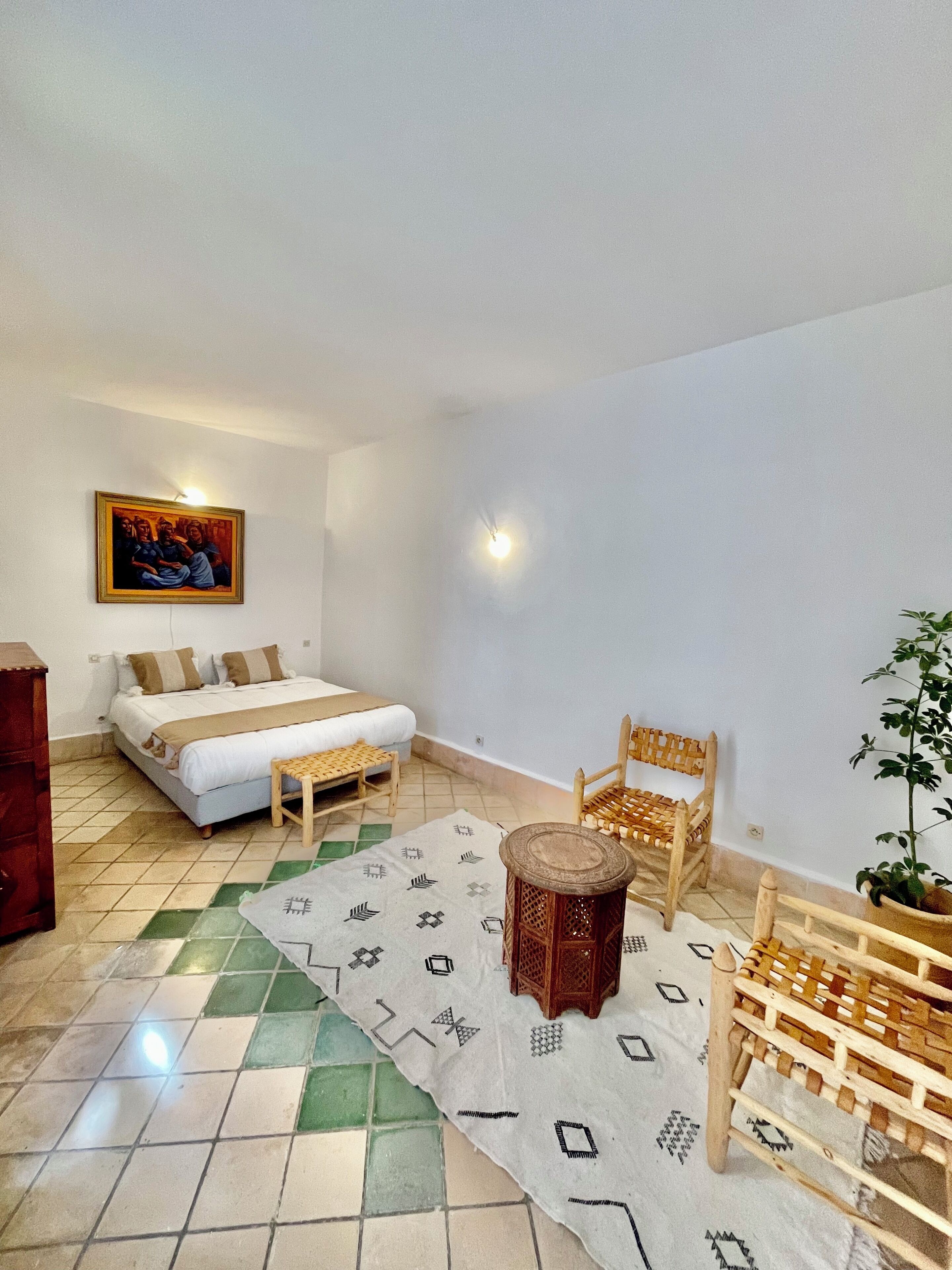 A room with a bed, a wooden chair, a small table, a potted plant, and a painting on the wall.
