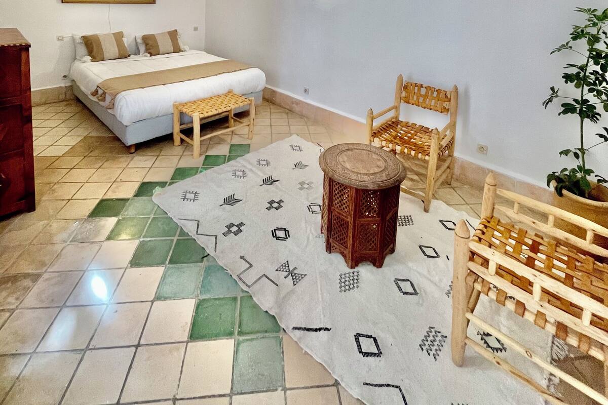 Una habitación con una cama, una silla de madera, una mesita, una planta en maceta y un cuadro en la pared.