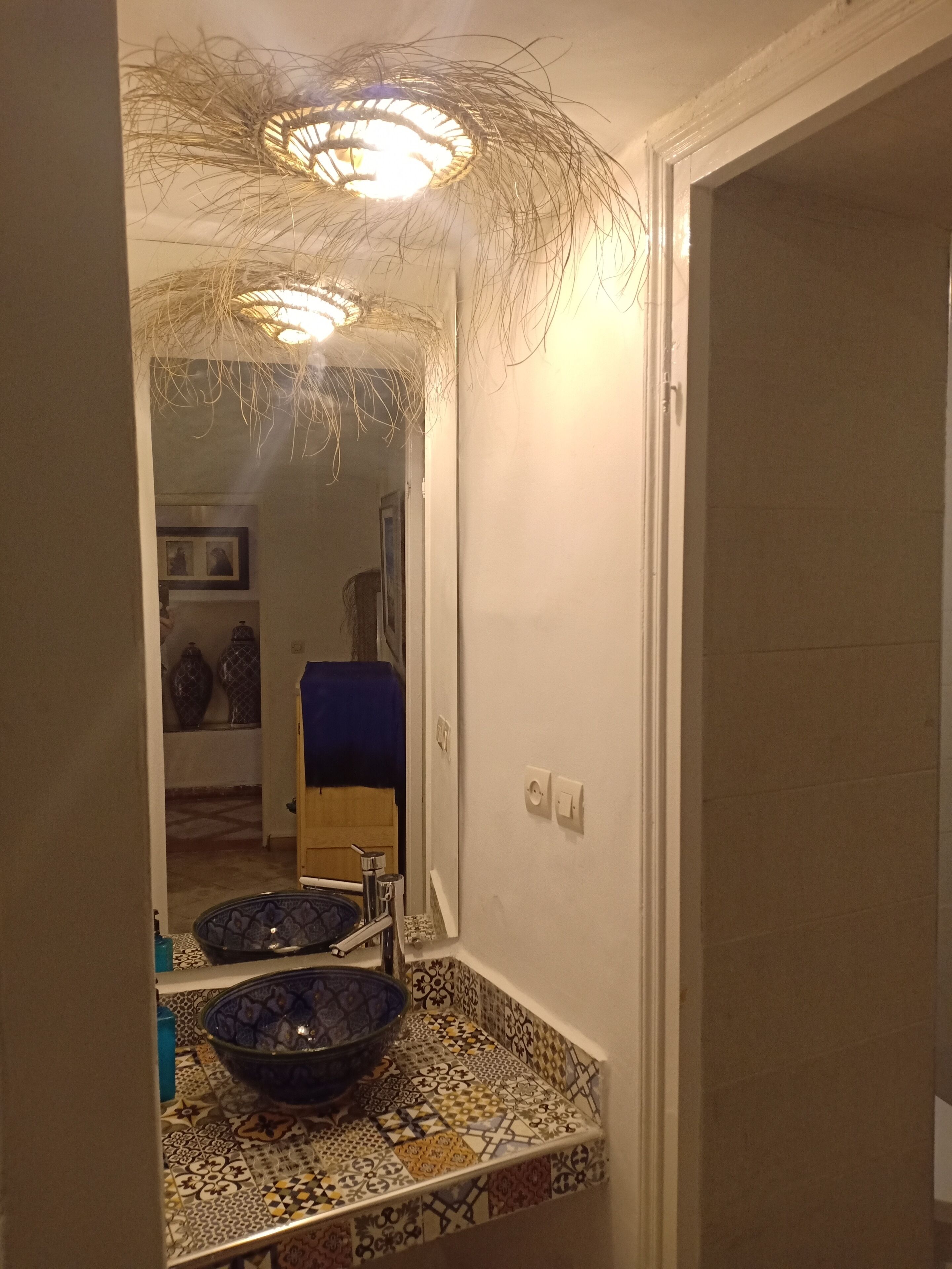 A bathroom with a large mirror, a blue decorative bowl, patterned tiles, and a ceiling light fixture.