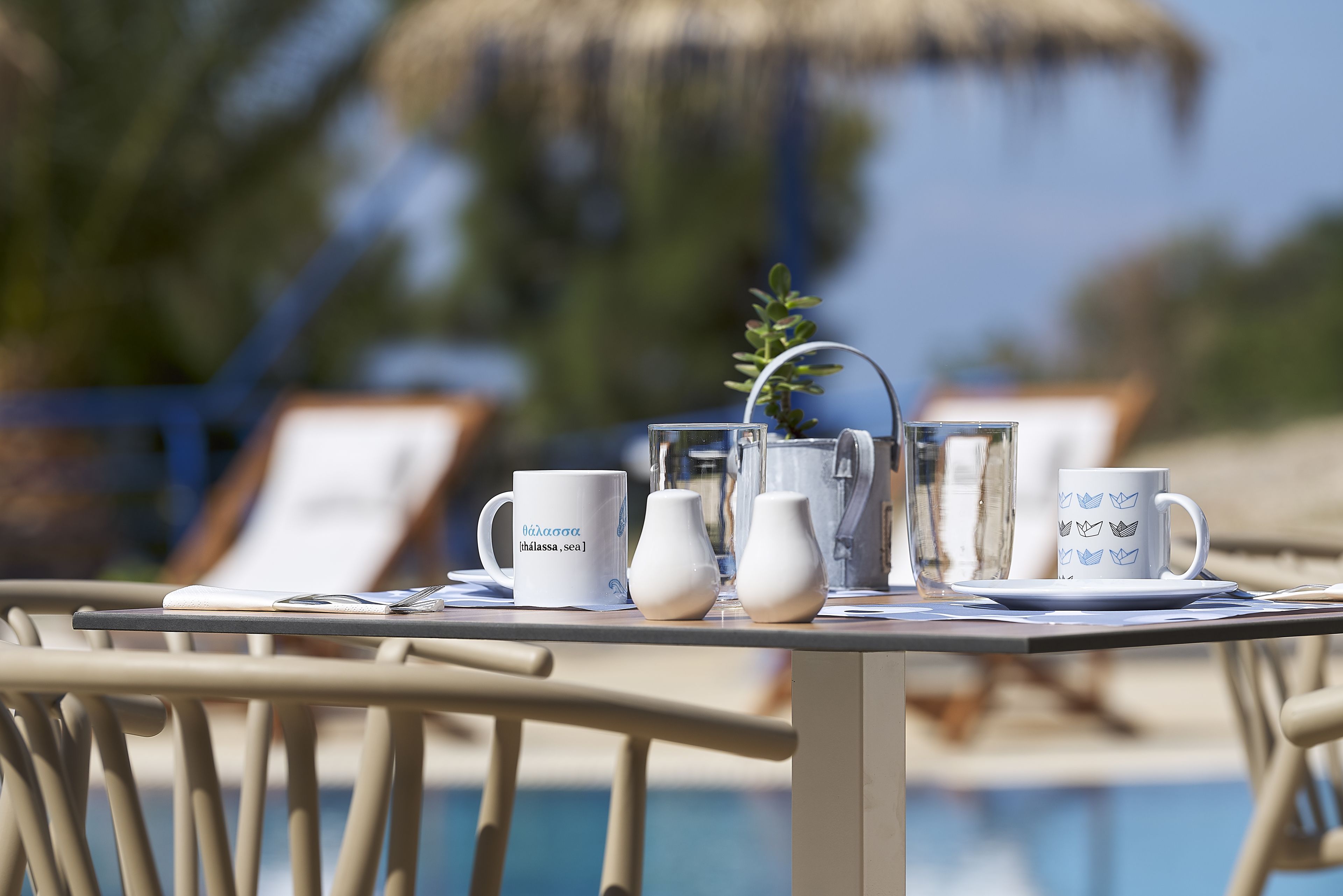 A table with a white mug, a white pitcher, and a white cup with a blue design, set on a table near a pool.