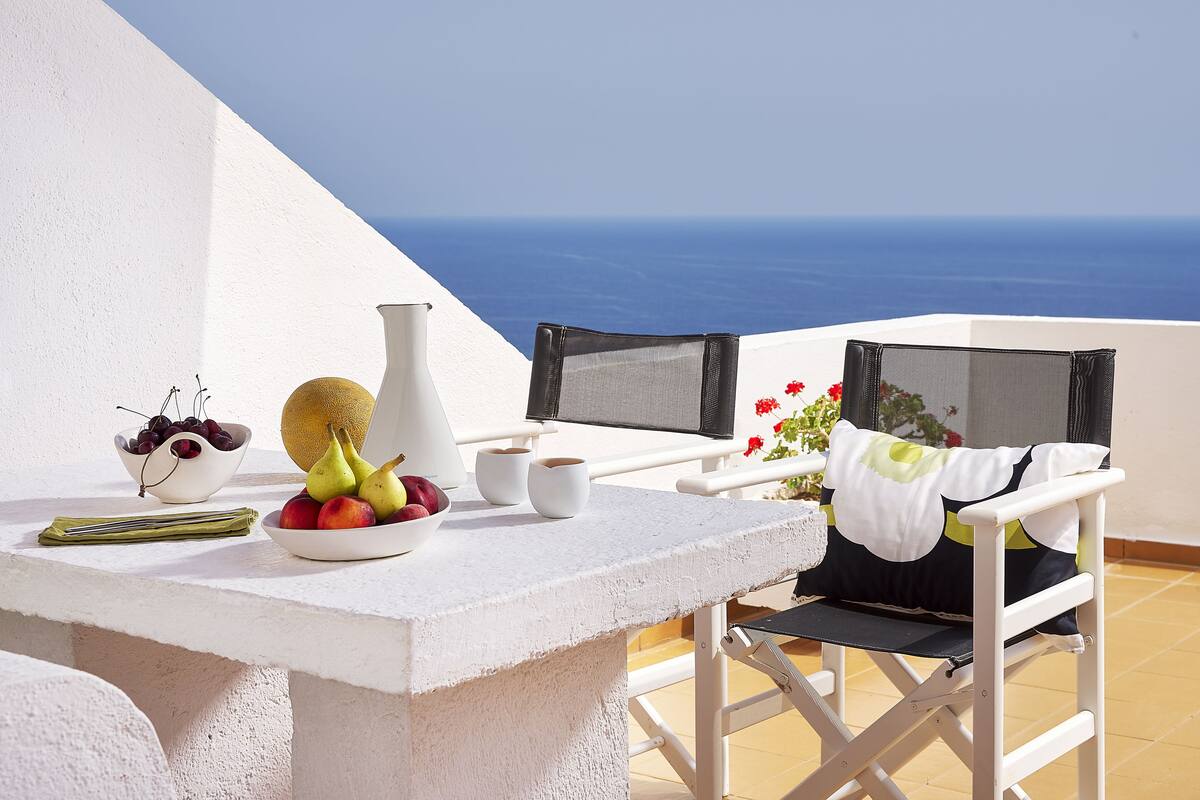 A terrace with a table set for two, featuring a bowl of fruit, a vase, and a cup. A chair with a cushion is positioned facing the terrace.