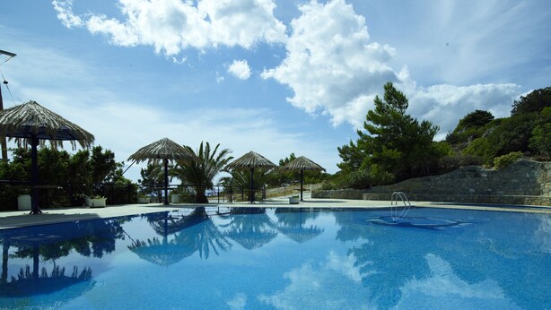 A swimming pool with clear blue water, surrounded by palm trees and thatched umbrellas. - Nakou Village (Ierapetra)