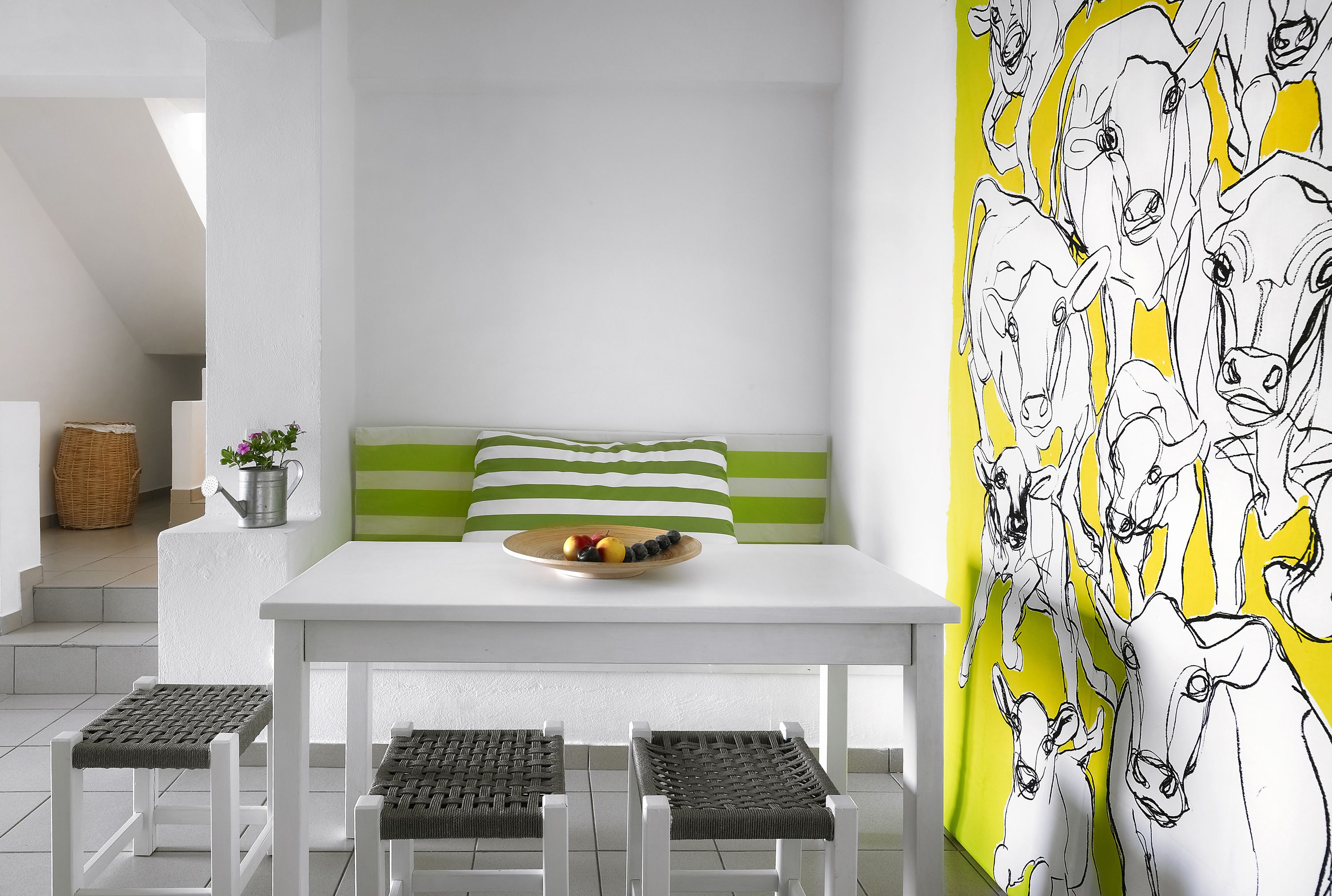 Un intérieur moderne avec une table blanche, des coussins à rayures vertes, des chaises en osier et un mur orné d’un motif de vaches.