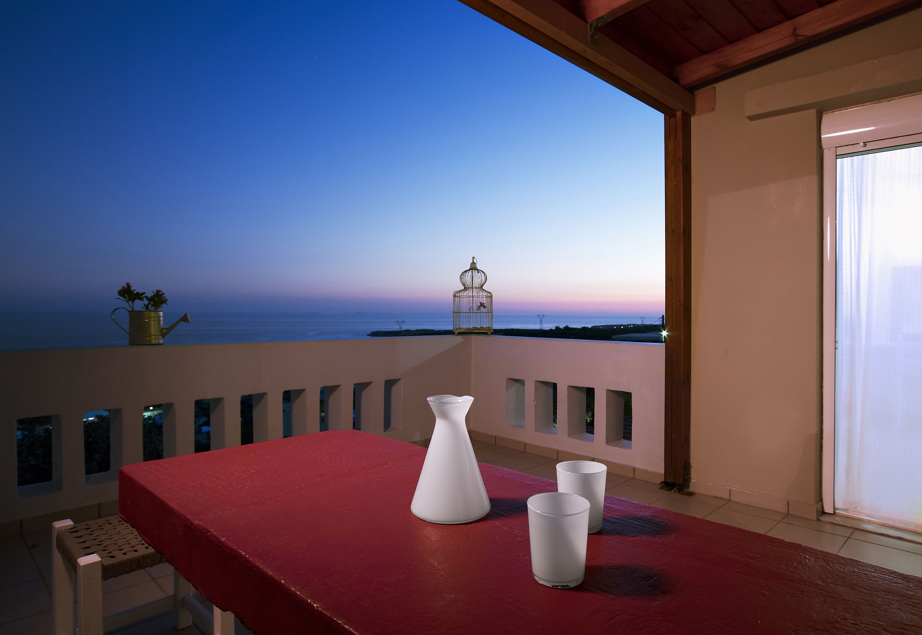 A balcony with a red table, a white vase, and two glasses.
