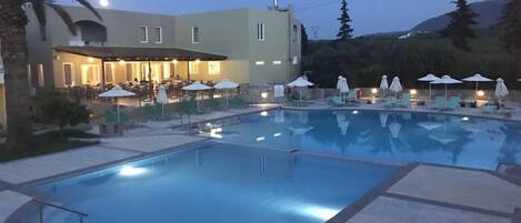 Un hotel con piscina, mobiliario de exterior y montañas al fondo.