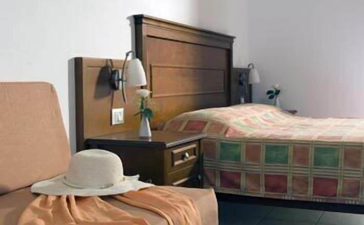 A bedroom with a wooden headboard, a bedside table with lamps, a bed with a colorful quilt, and a white hat on the bed.