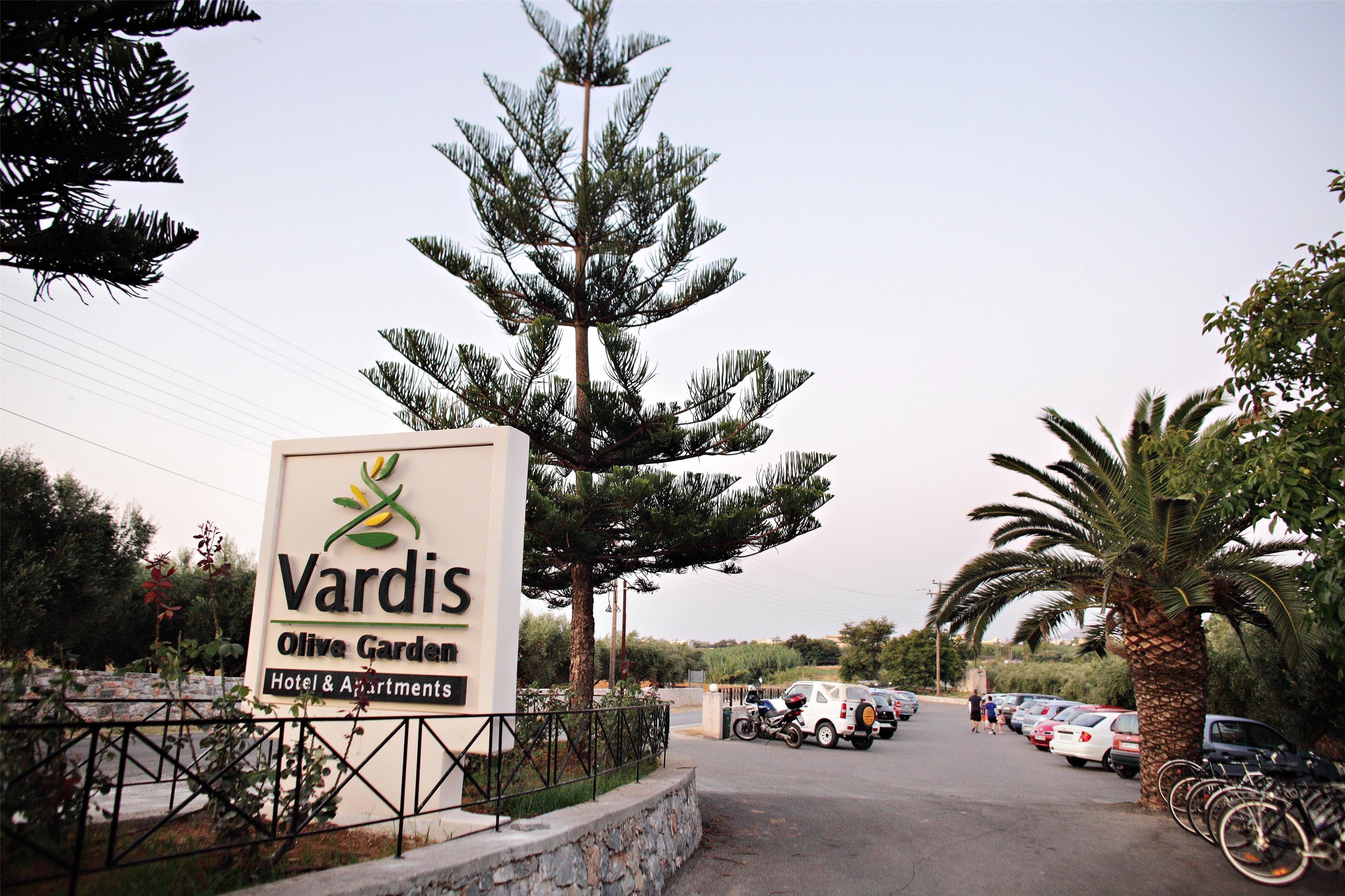 A sign for Vardis Olive Garden Hotel & Apartments with a parking lot and parked cars.