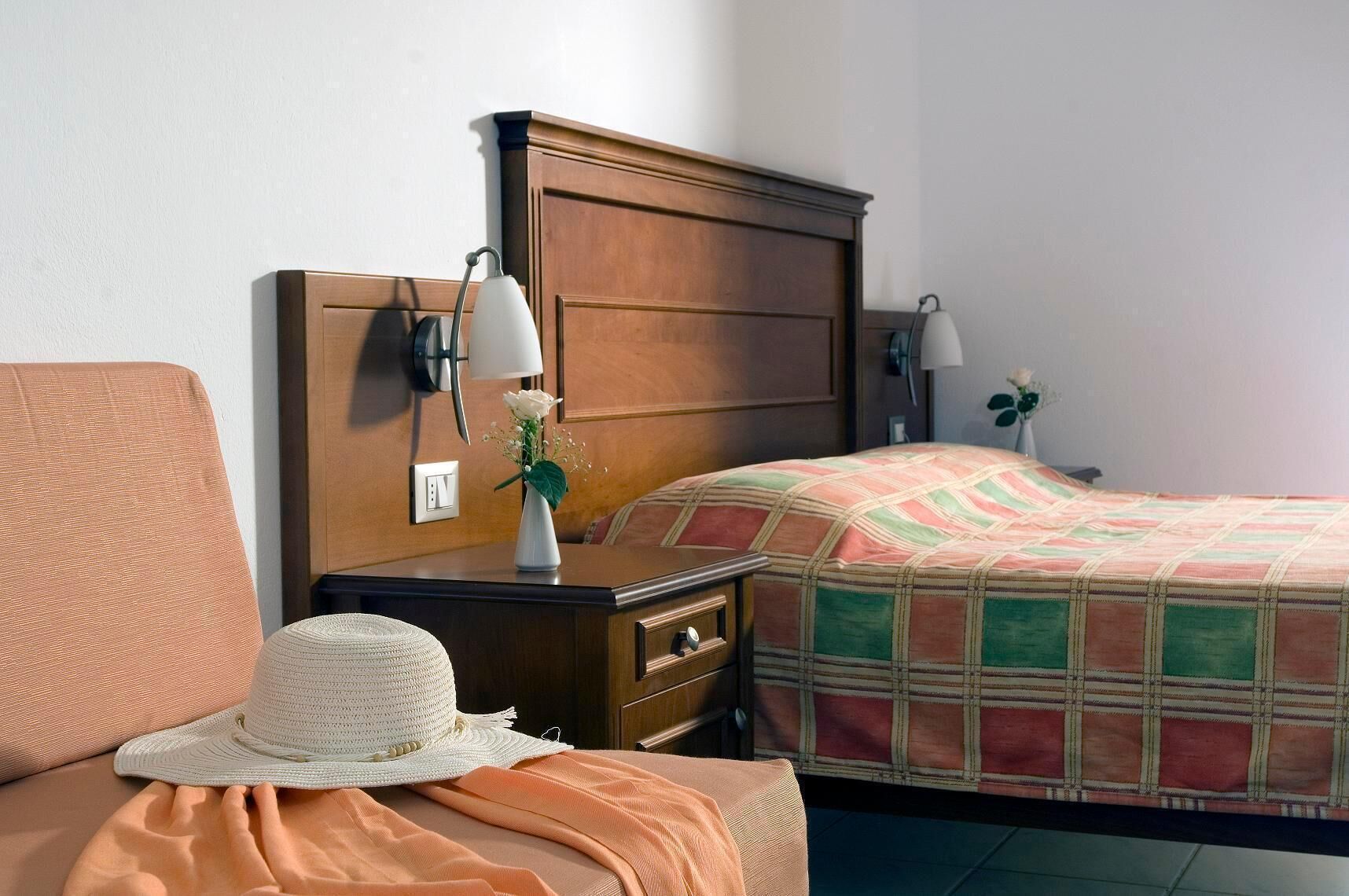 Une chambre d’hôtel avec une tête de lit en bois, un lit recouvert d’une couette à carreaux, une table de chevet avec une lampe, un vase rempli de fleurs et un panier en osier.