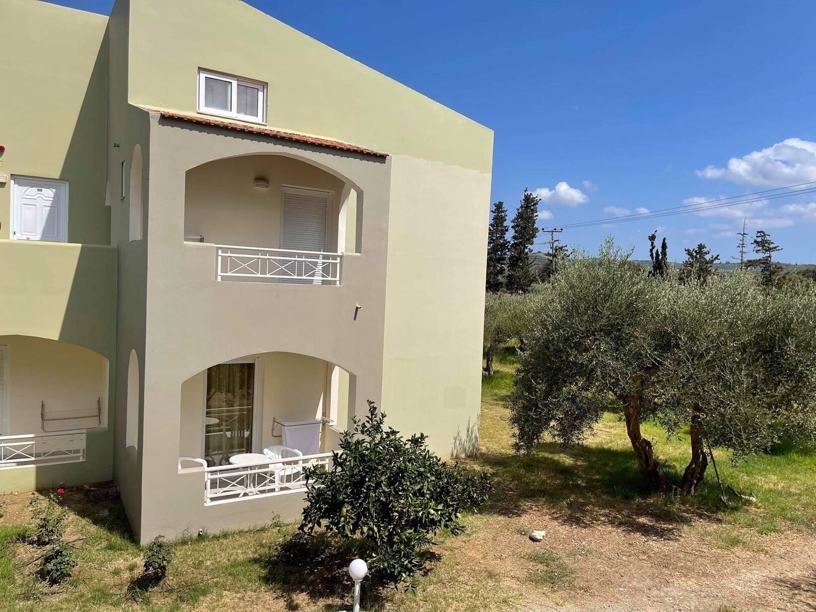 Un edificio de dos pisos con balcón, asientos al aire libre y olivos en el fondo.