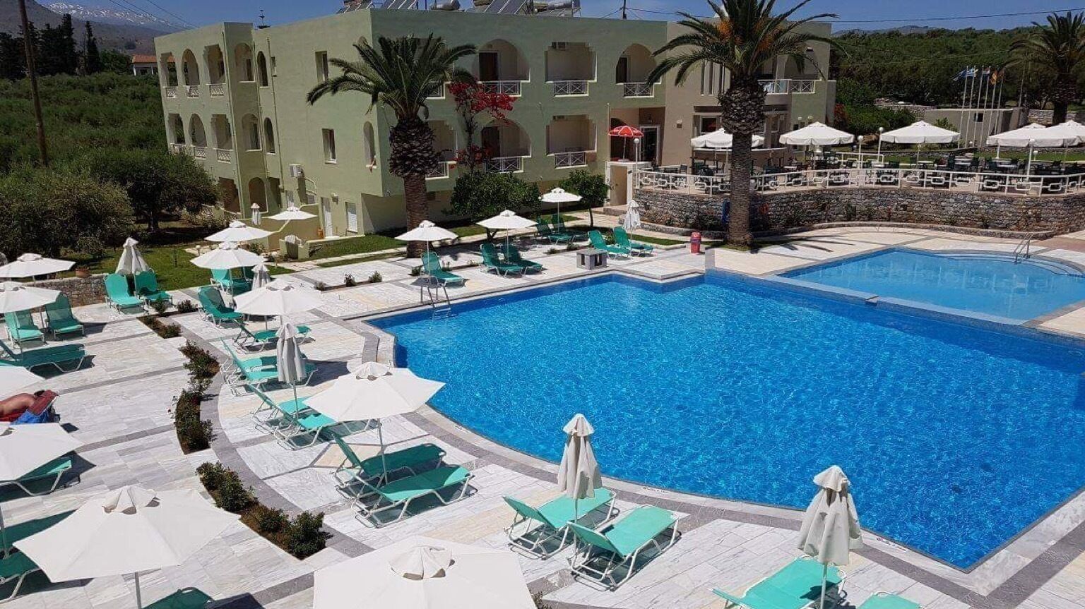Un área de piscina con múltiples sillas de descanso y sombrillas, una piscina grande y un edificio con balcones.