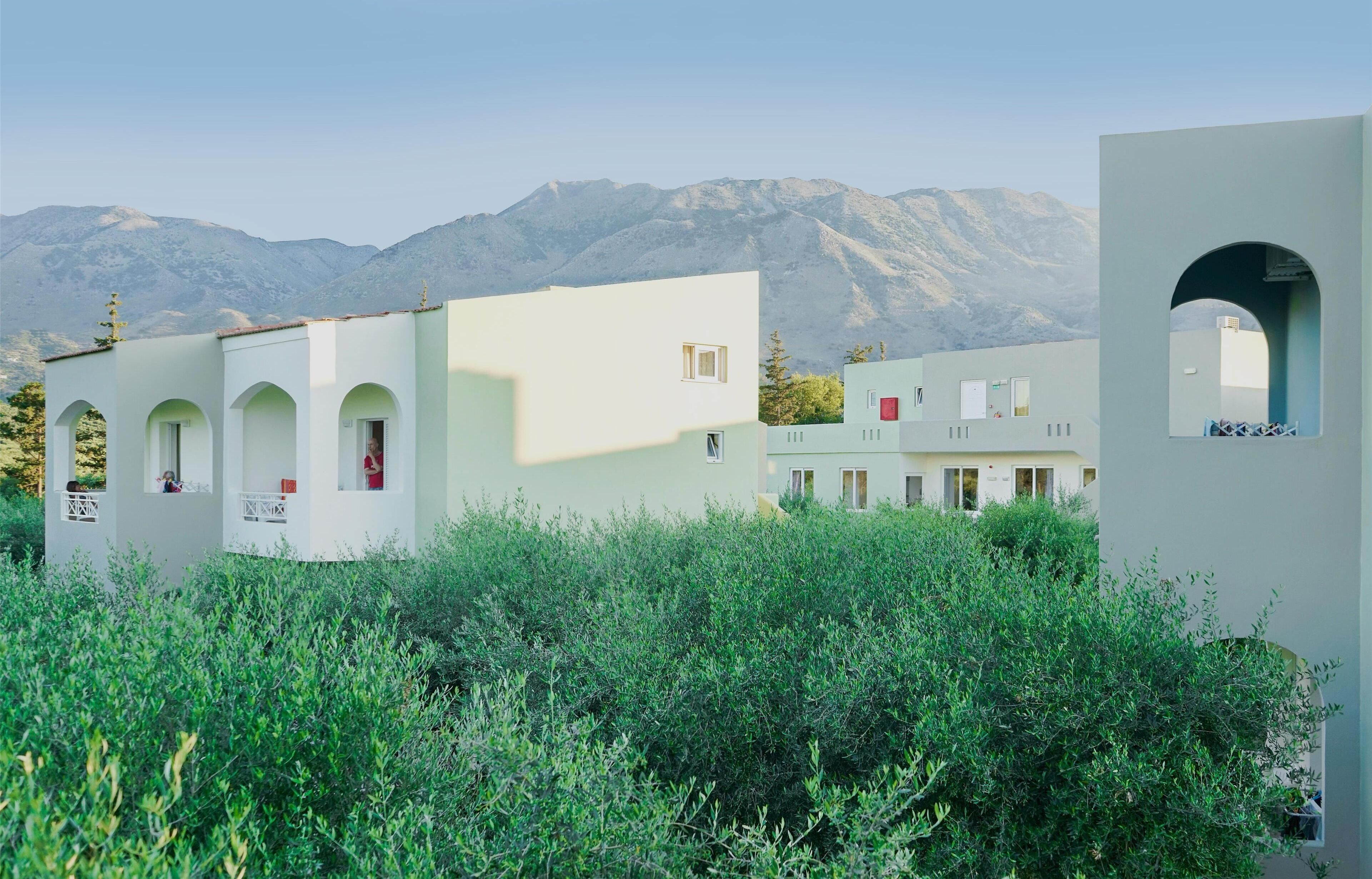 A modern building with arched windows and a flat roof, surrounded by dense greenery and mountains in the background.