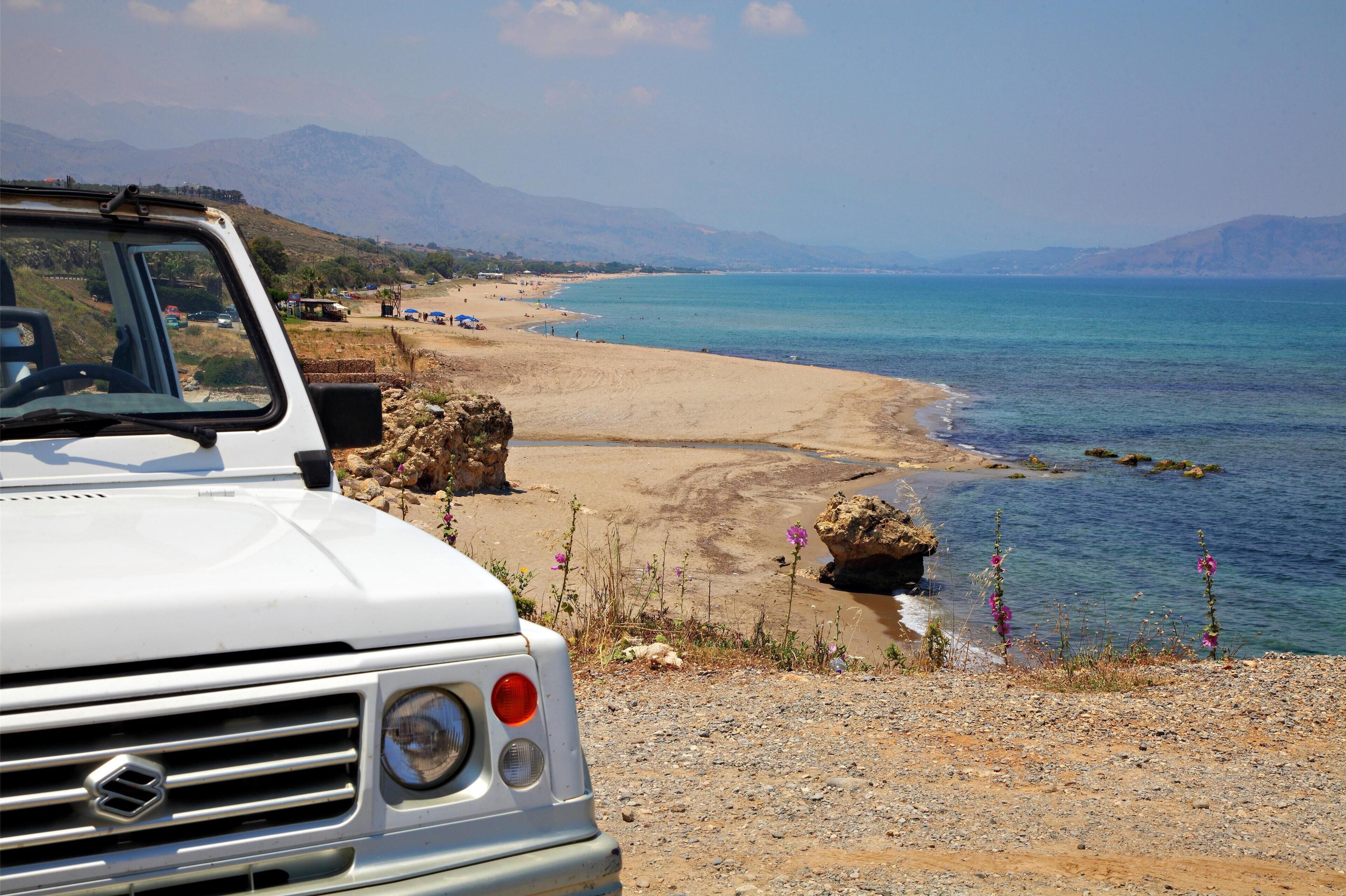 Un vehículo blanco estacionado en una playa rocosa con vista despejada al mar y montañas distantes.