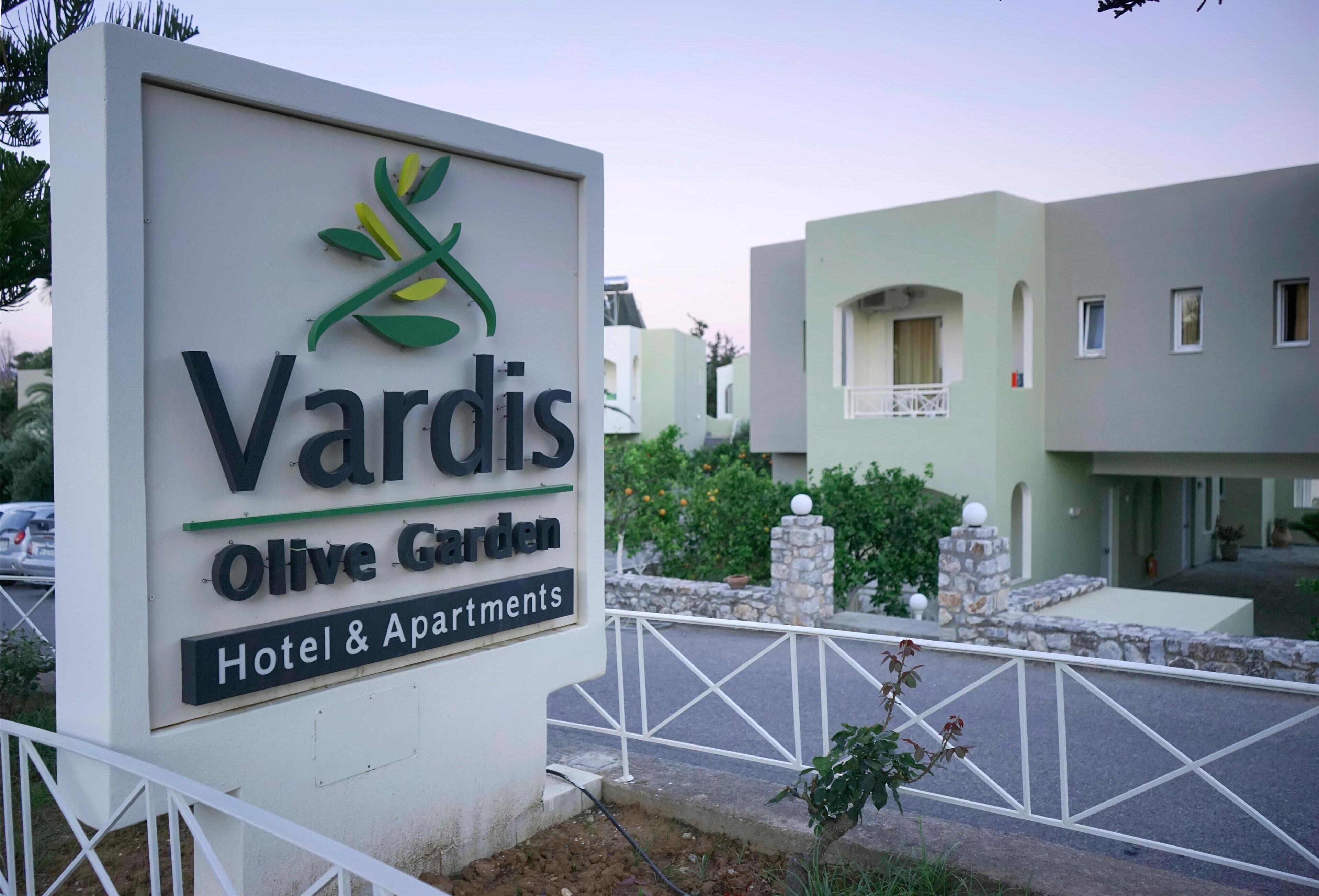 A sign for Vardis Olive Garden Hotel & Apartments with a modern building in the background.