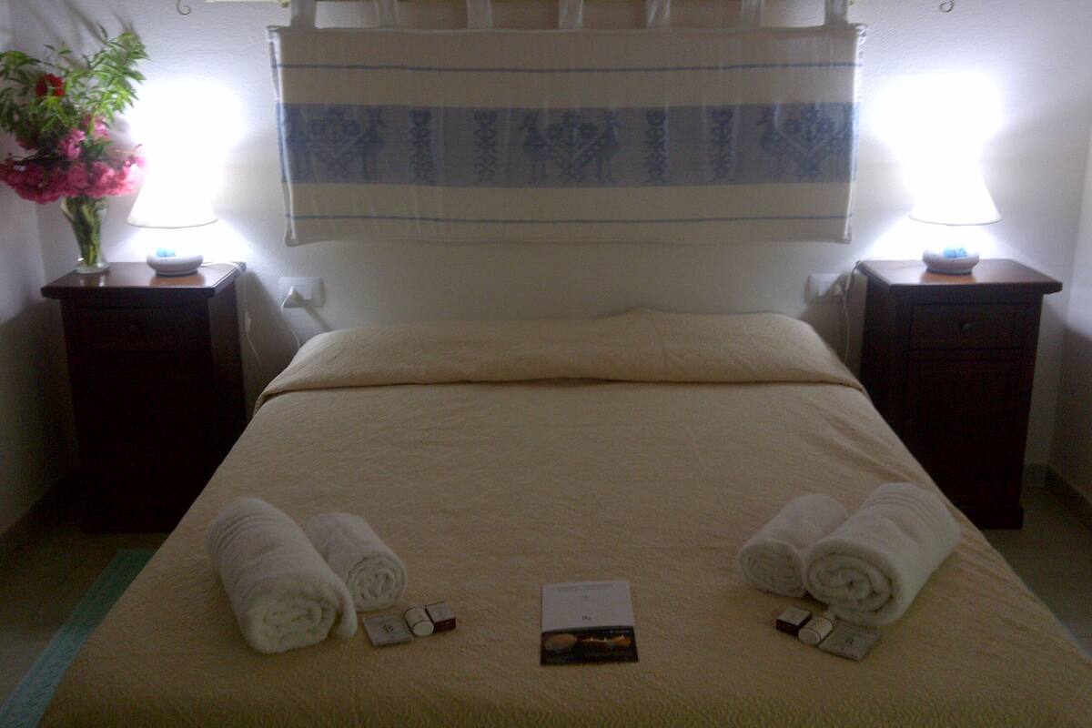A neatly made bed with towels and toiletries, two bedside tables with lamps, and a floral arrangement on one table.
