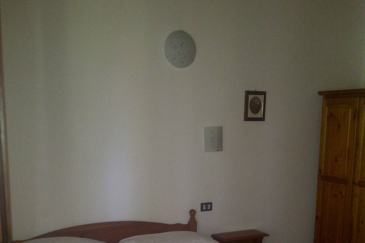 A single bed with a floral bedspread and two rolled towels, a wooden nightstand, a wooden wardrobe, and a wall-mounted light switch.