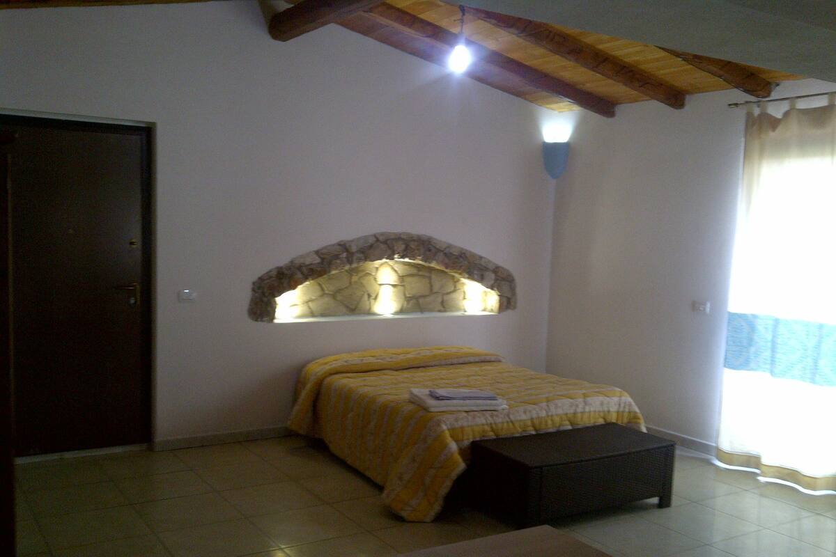 A bedroom with a bed, a bench, a stone arch window, and a door.
