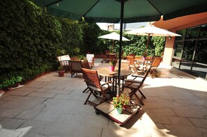 Un patio al aire libre con mesas y sillas de madera, cubierto por sombrillas, rodeado de exuberante follaje.