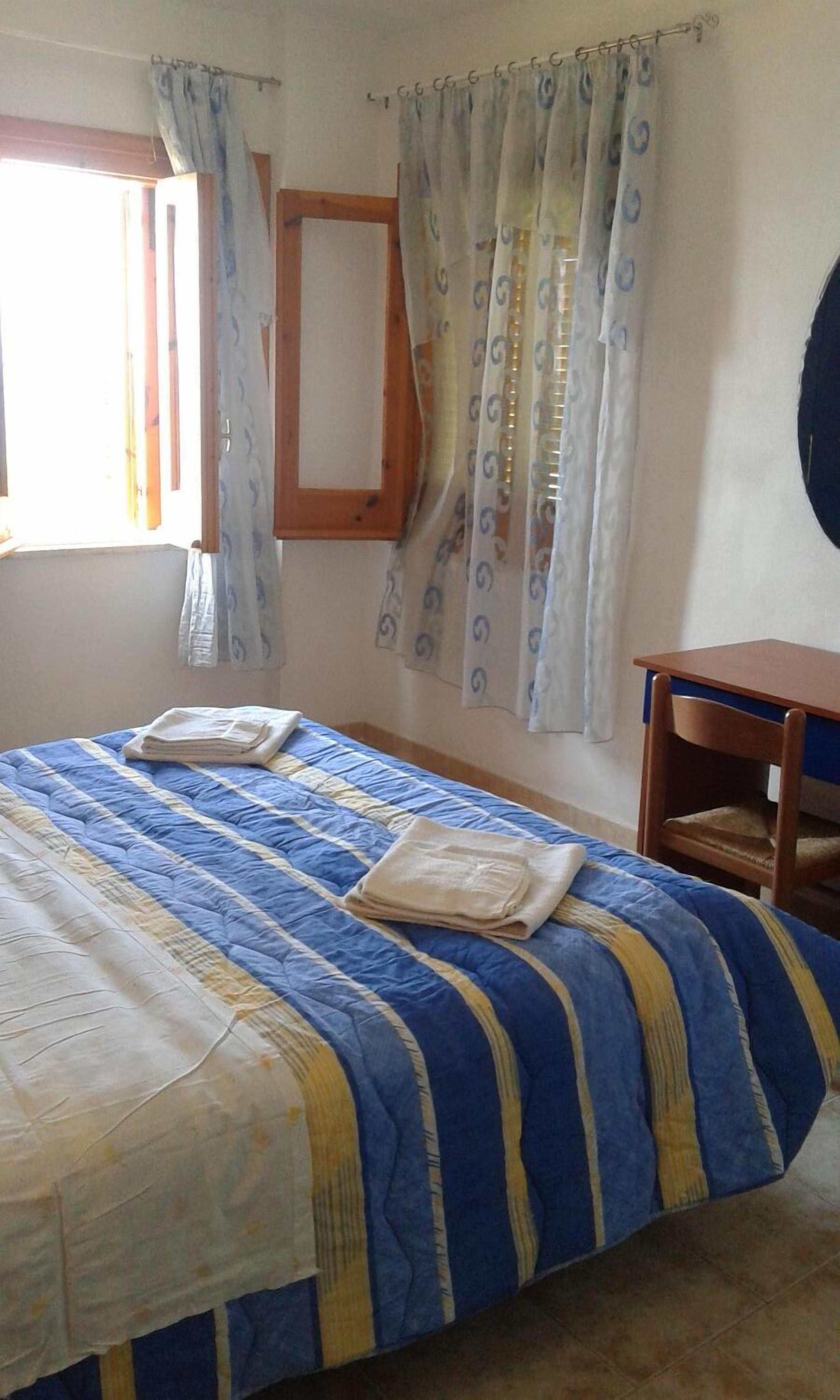Quarto com cama, cadeira de madeira, mesa pequena e janela com cortinas.