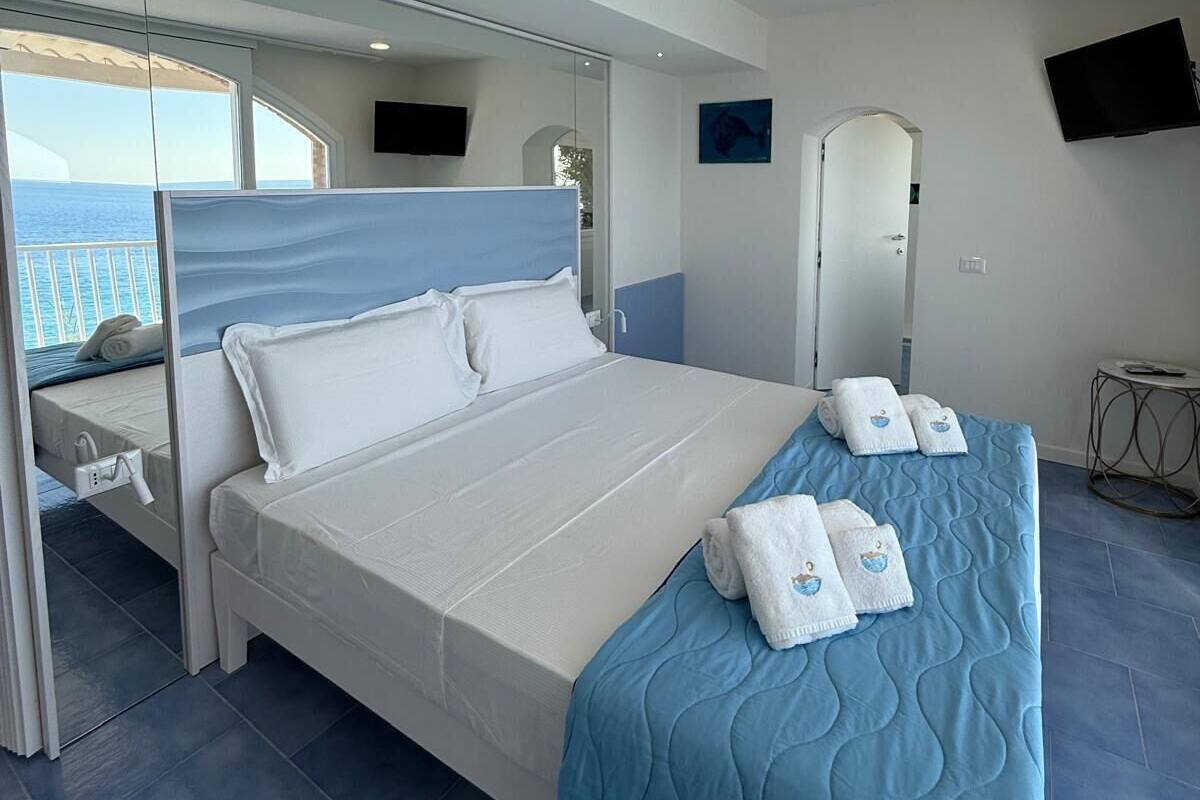 A modern hotel room with a large bed, a view of the sea, a flat-screen TV, and a small table.