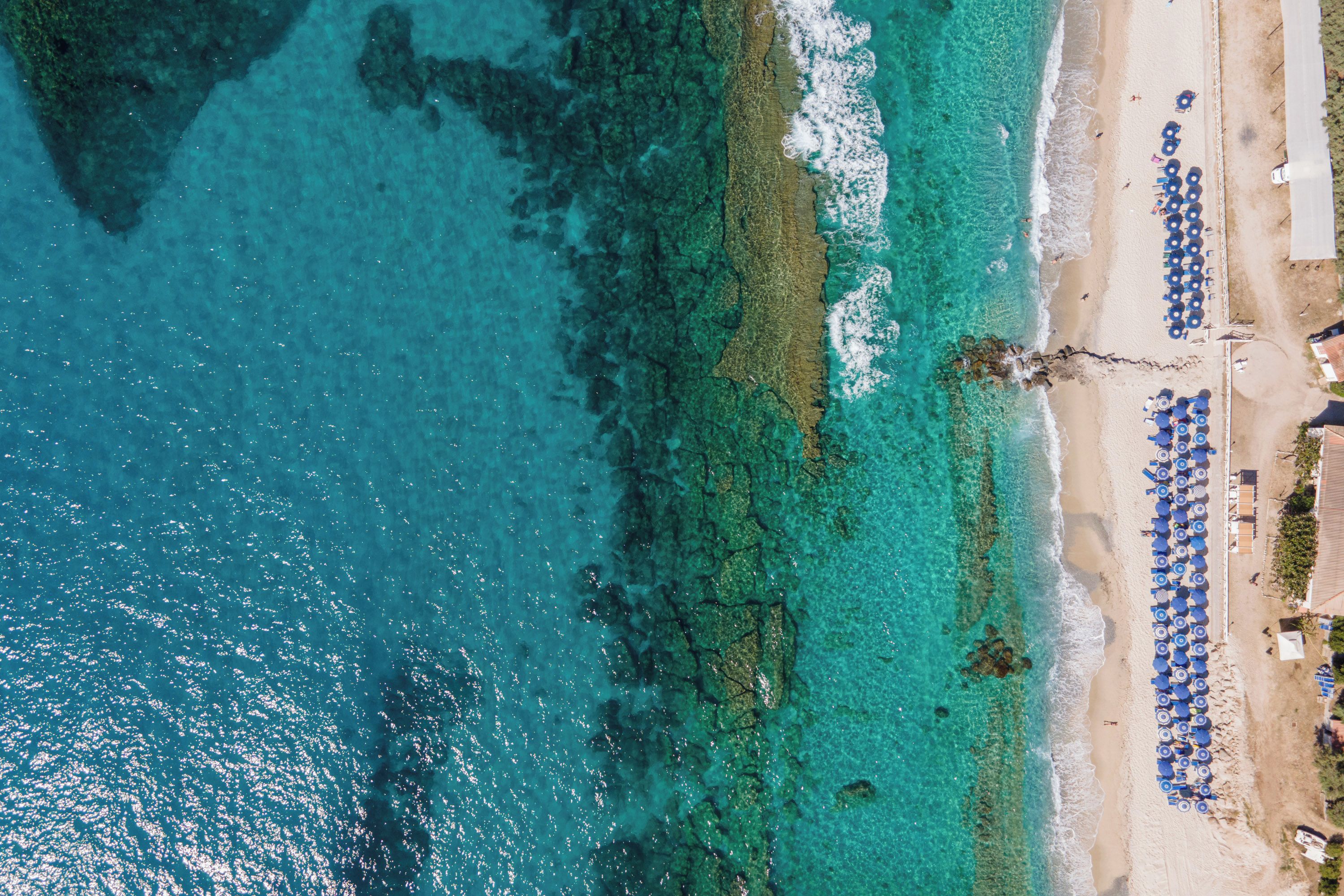 An aerial view of a beach with clear turquoise waters, a sandy shore, and rows of blue umbrellas.