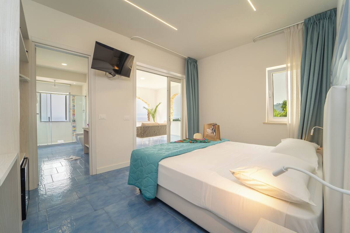 A hotel room with a bed, a TV mounted on the wall, a window with curtains, and a view of the outside.