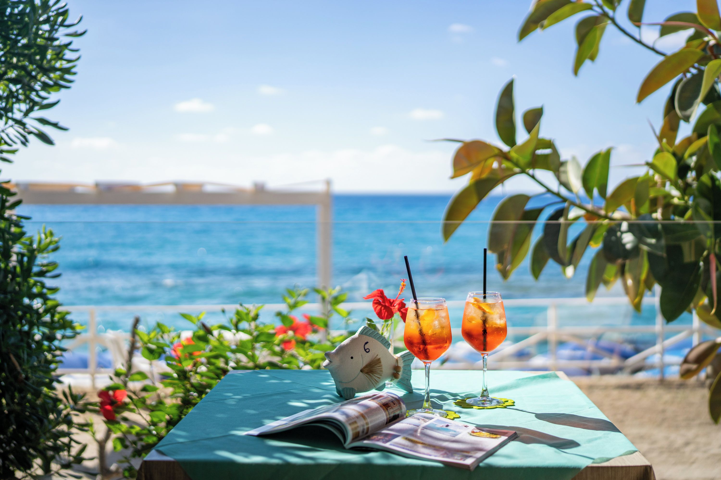 A table with drinks, a book, and a decorative item on a beach overlooking the sea.