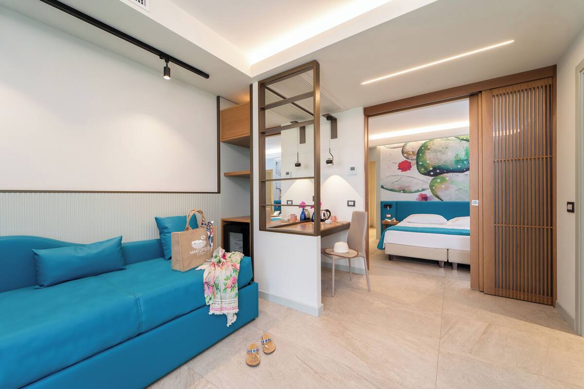 A modern hotel room with a blue sofa, a desk with a mirror, a bed with a colorful mural, and a sliding wooden door.
