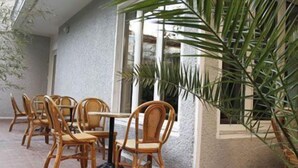 Un espacio al aire libre con sillas y mesas de madera, rodeado de plantas y la fachada de un edificio.