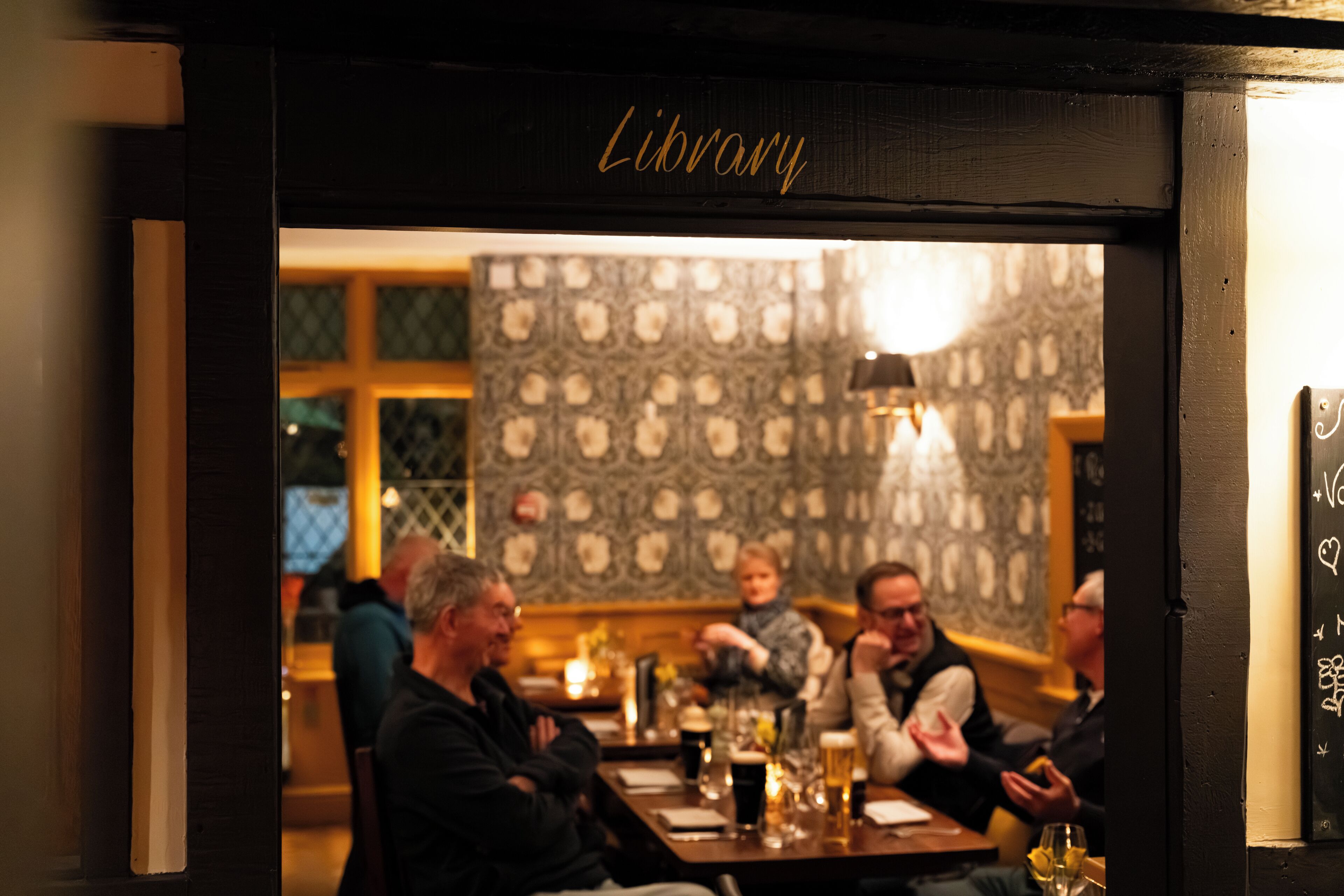Een restaurant interieur met een 'Library' bord, behang met patroon en gasten die aan tafels zitten.