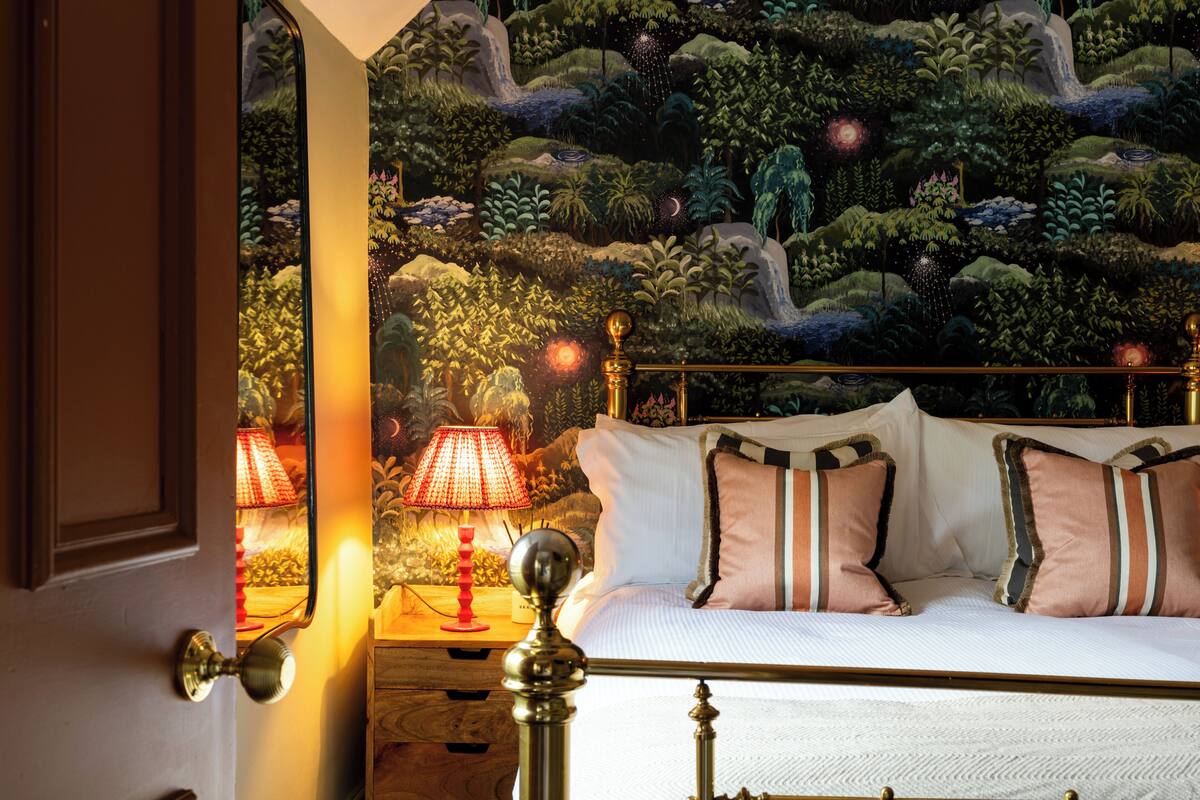 Quarto com cama, almofadas, mesa de cabeceira, abajur e um mural na parede.