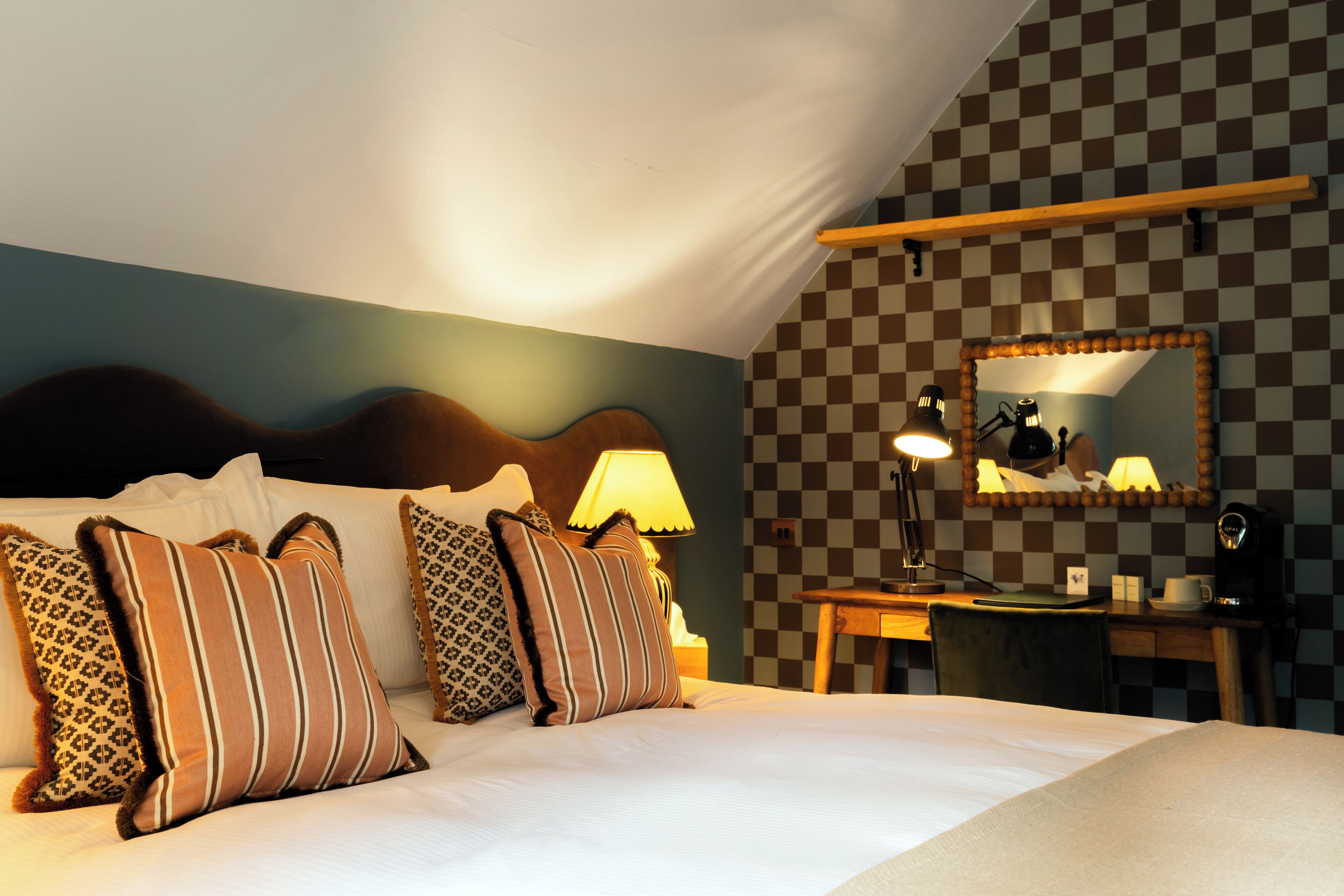 A hotel room with a bed, pillows, a nightstand, a desk, a mirror, and a lamp.