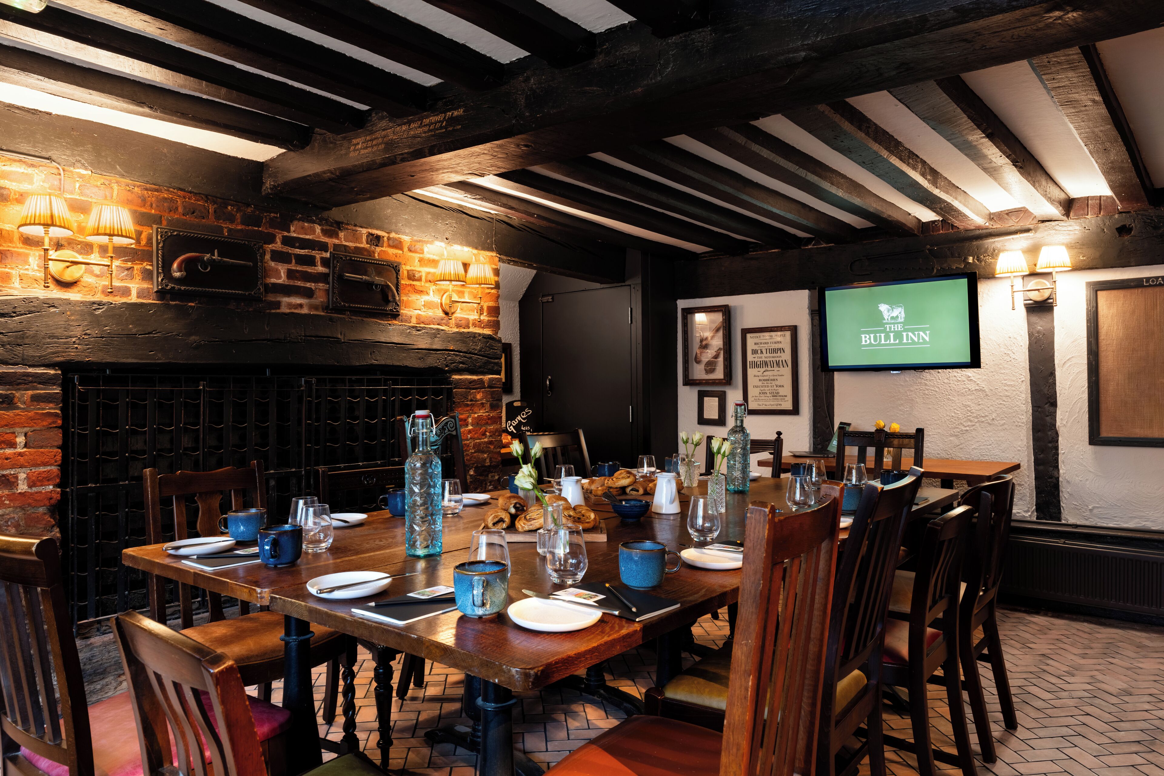Egy étkező, melynek egyik fala téglából van, a mennyezetén pedig fagerendák találhatók, és egy televízión a „The Bull Inn” logója látható.