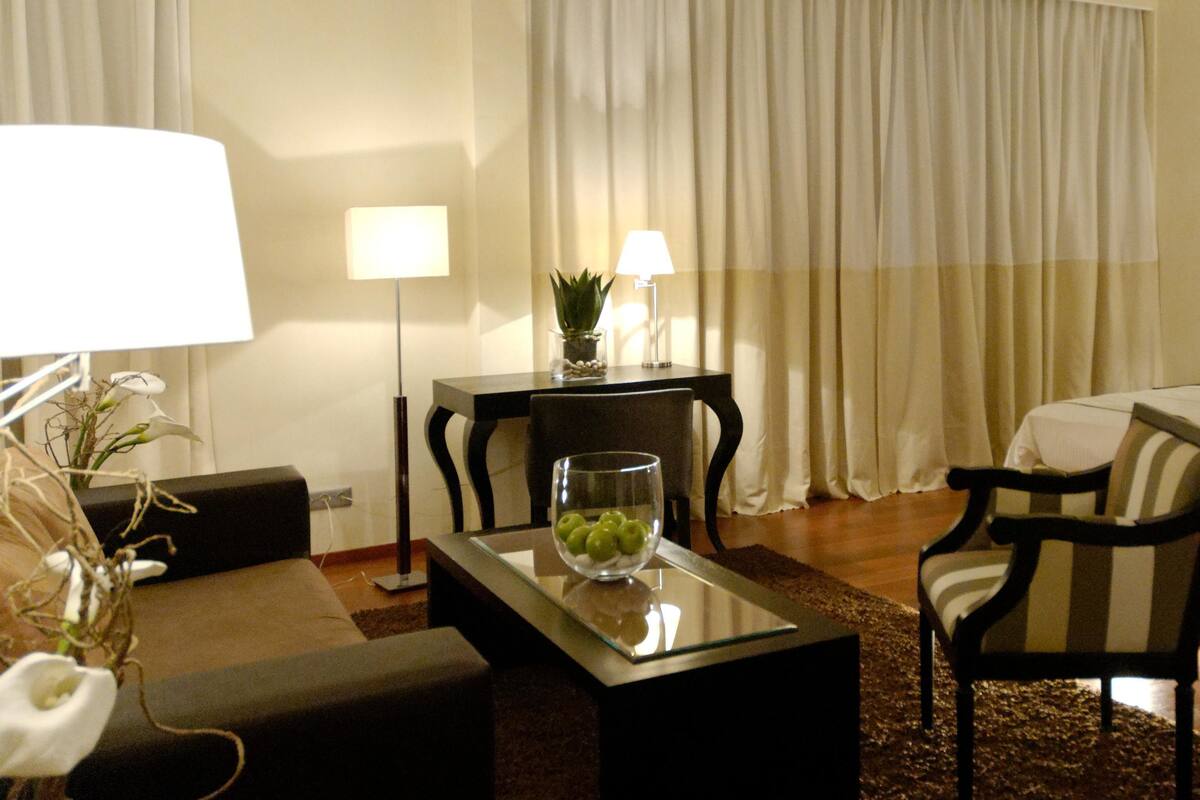 Een hotelkamer met een bed, een bureau met lamp, een stoel en een bijzettafeltje met een schaal fruit.