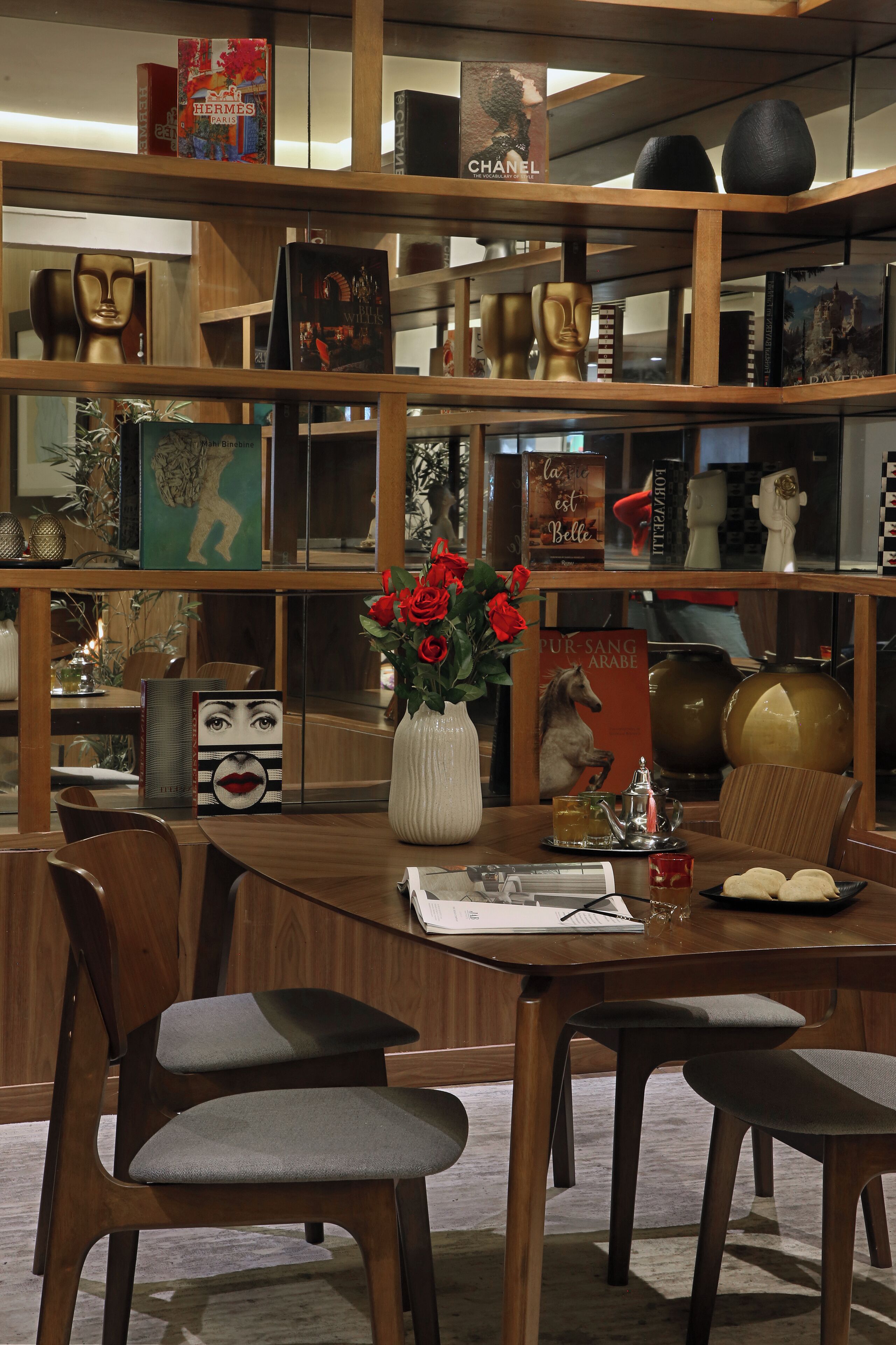 Un intérieur moderne avec une table à manger en bois, des chaises et une bibliothèque remplie de livres et d’objets décoratifs.