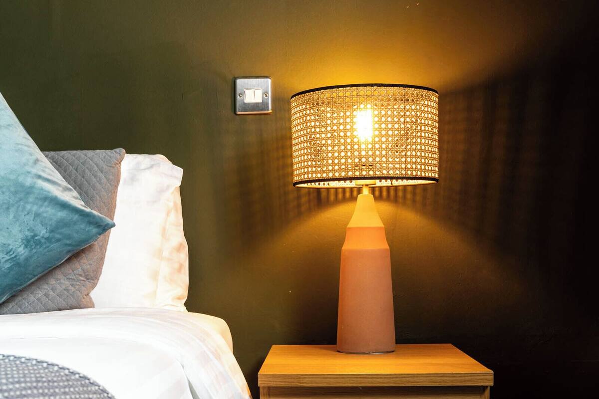 Una cama bien hecha con edredón blanco y almohadas, una mesita de noche de madera con una lámpara e interruptor de pared.
