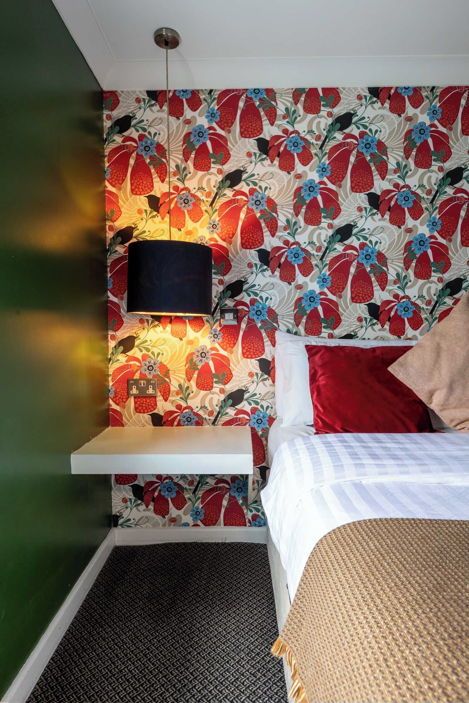 A bedroom with a patterned wallpaper, a hanging lamp, a bed with red and white bedding, and a small bedside table.