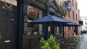 L’entrée d’un hôtel, avec une enseigne, des tables et des chaises à l’extérieur, protégées par des parasols, et des plantes en pot.
