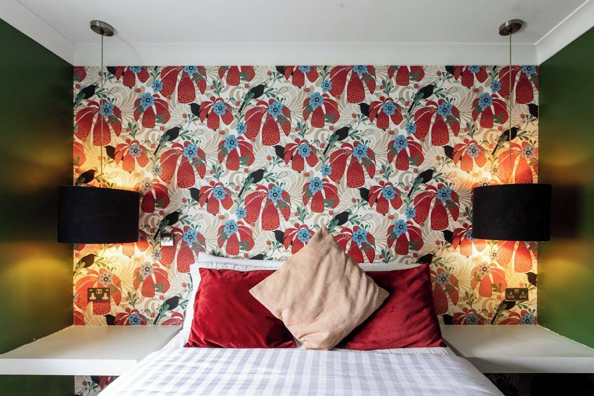 Un dormitorio con papel tapiz floral, una cama con almohadas rojas y beige, y dos lámparas negras fijadas a la pared.