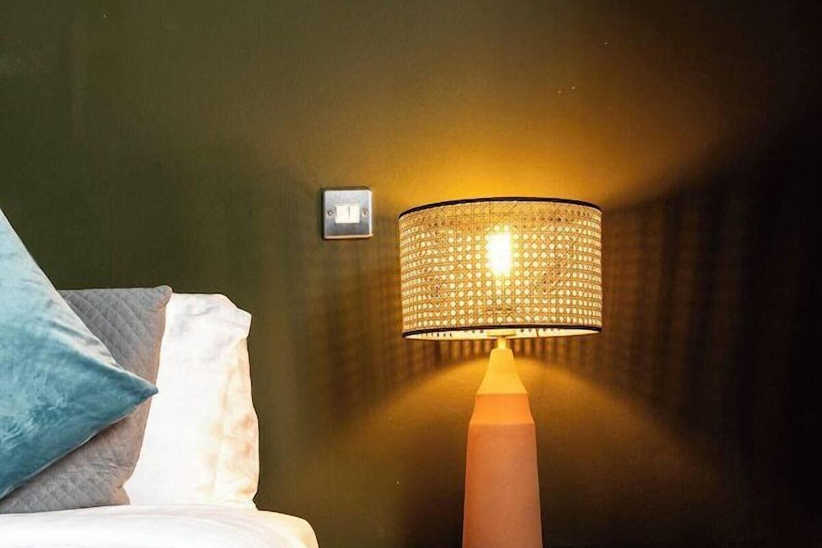 Una cama bien hecha con colcha blanca y gris estampada, una mesita de noche de madera con lámpara e interruptor de pared.