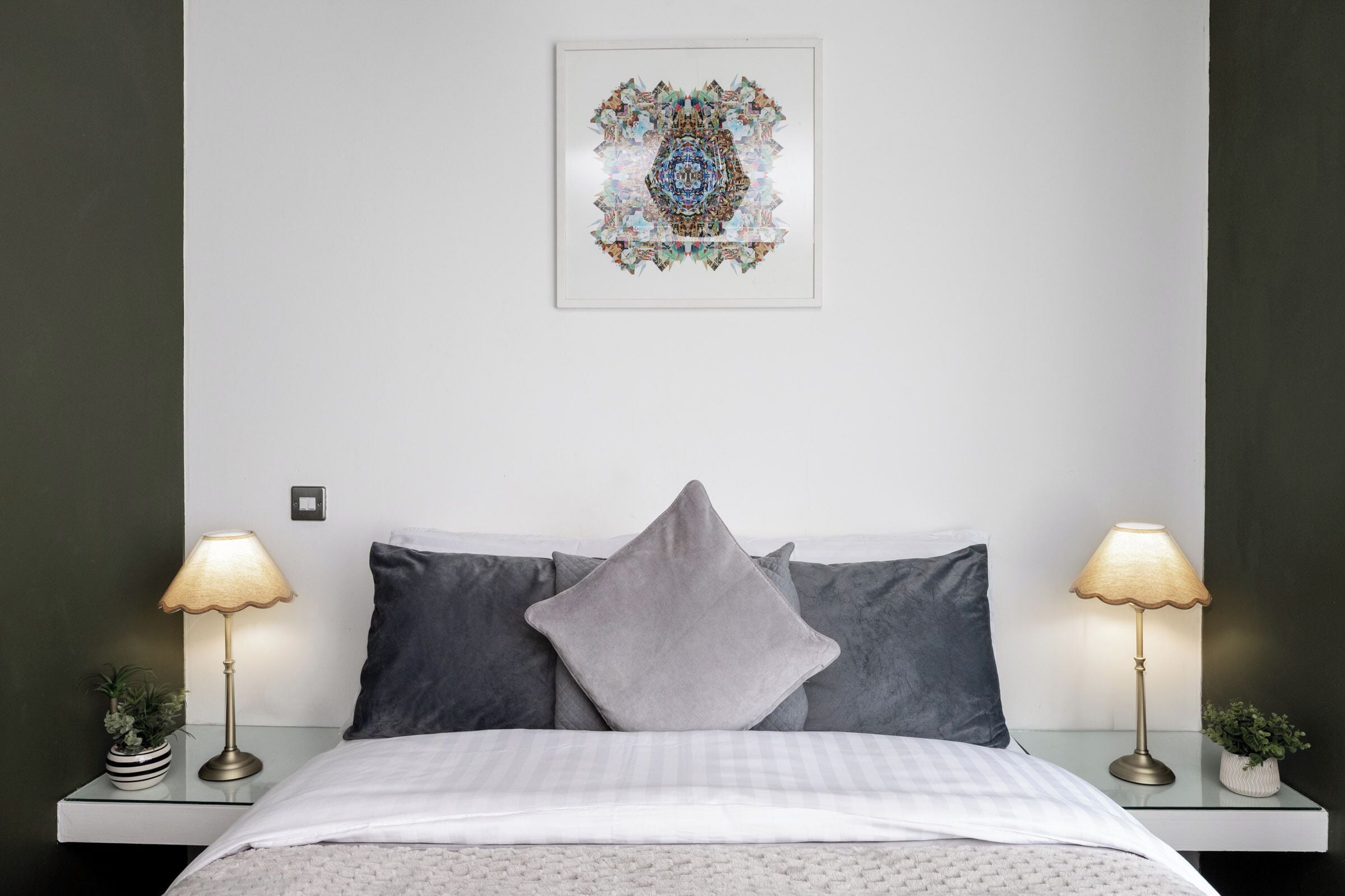 Una cama bien hecha con una colcha a cuadros, dos lámparas de noche y una obra enmarcada sobre la cama.