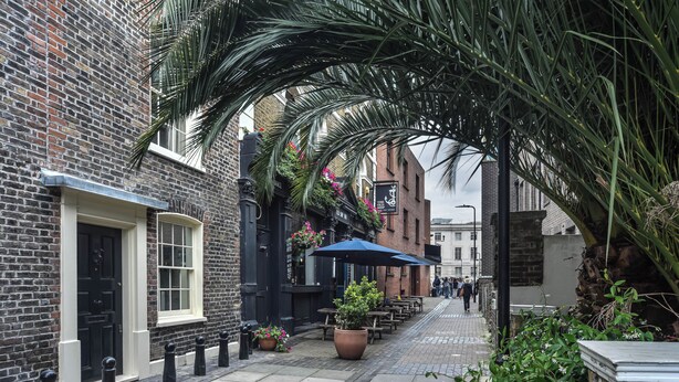 Une rue étroite bordée d’immeubles en briques, avec des terrasses et des plantes en pot.