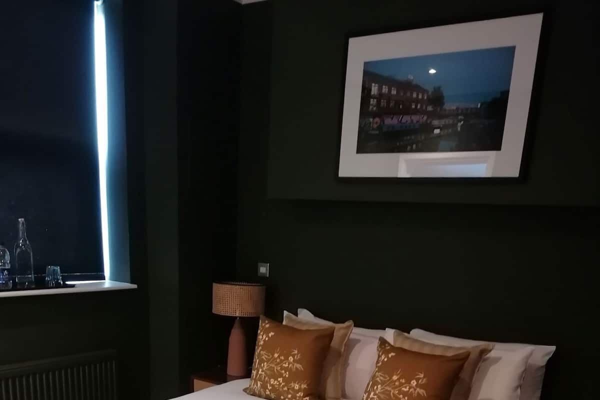 Una cama bien hecha con edredón blanco y dos almohadas marrones, una lámpara de noche, un cuadro enmarcado en la pared y una ventana con vistas a edificios.