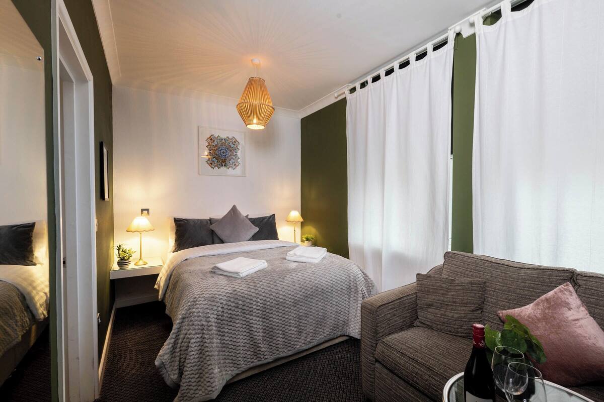 Una habitación de hotel con una cama, dos mesitas de noche con lámparas, un sofá, una mesa de vidrio con una botella y un vaso, y un elemento decorativo en la pared.