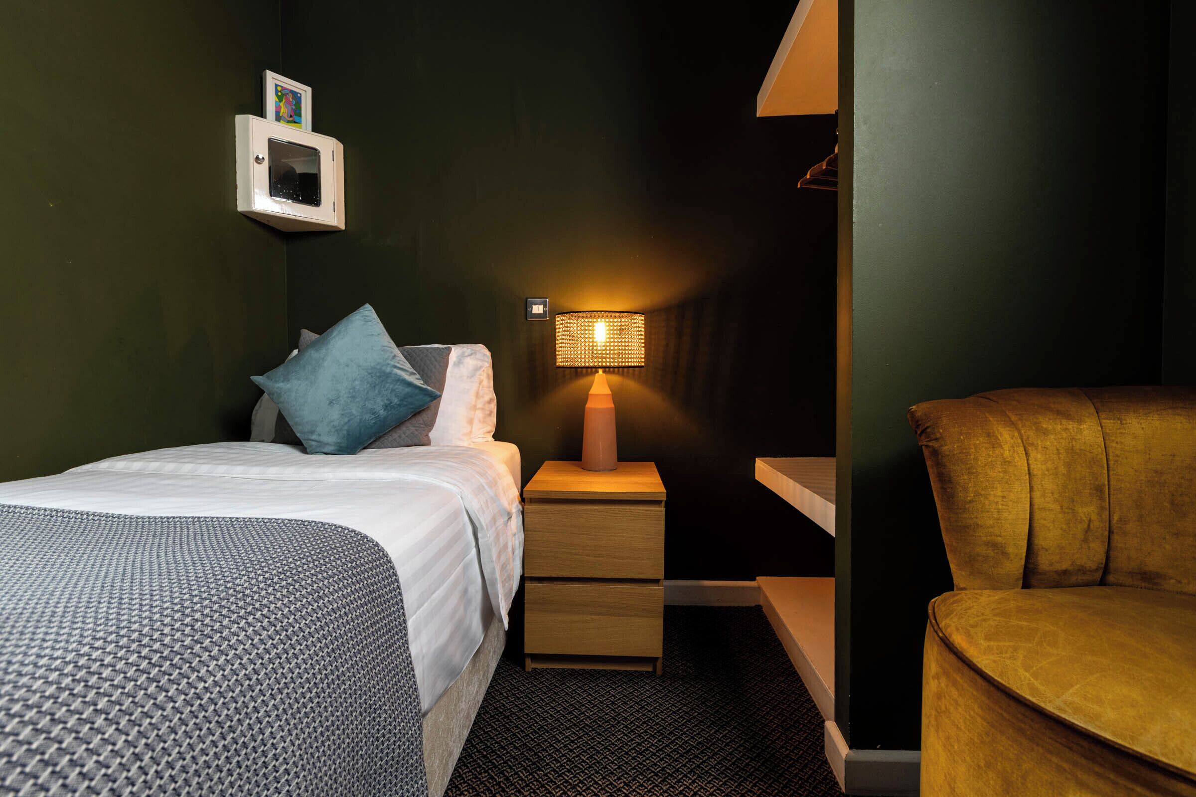 A hotel room with a bed, a nightstand with a lamp, a chair, and a small cabinet with a picture.